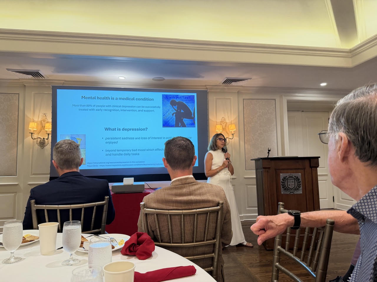 Karen B. Gerson speaking with a microphone to Plaza Club members seated at breakfast tables at Mission Hills Country Club